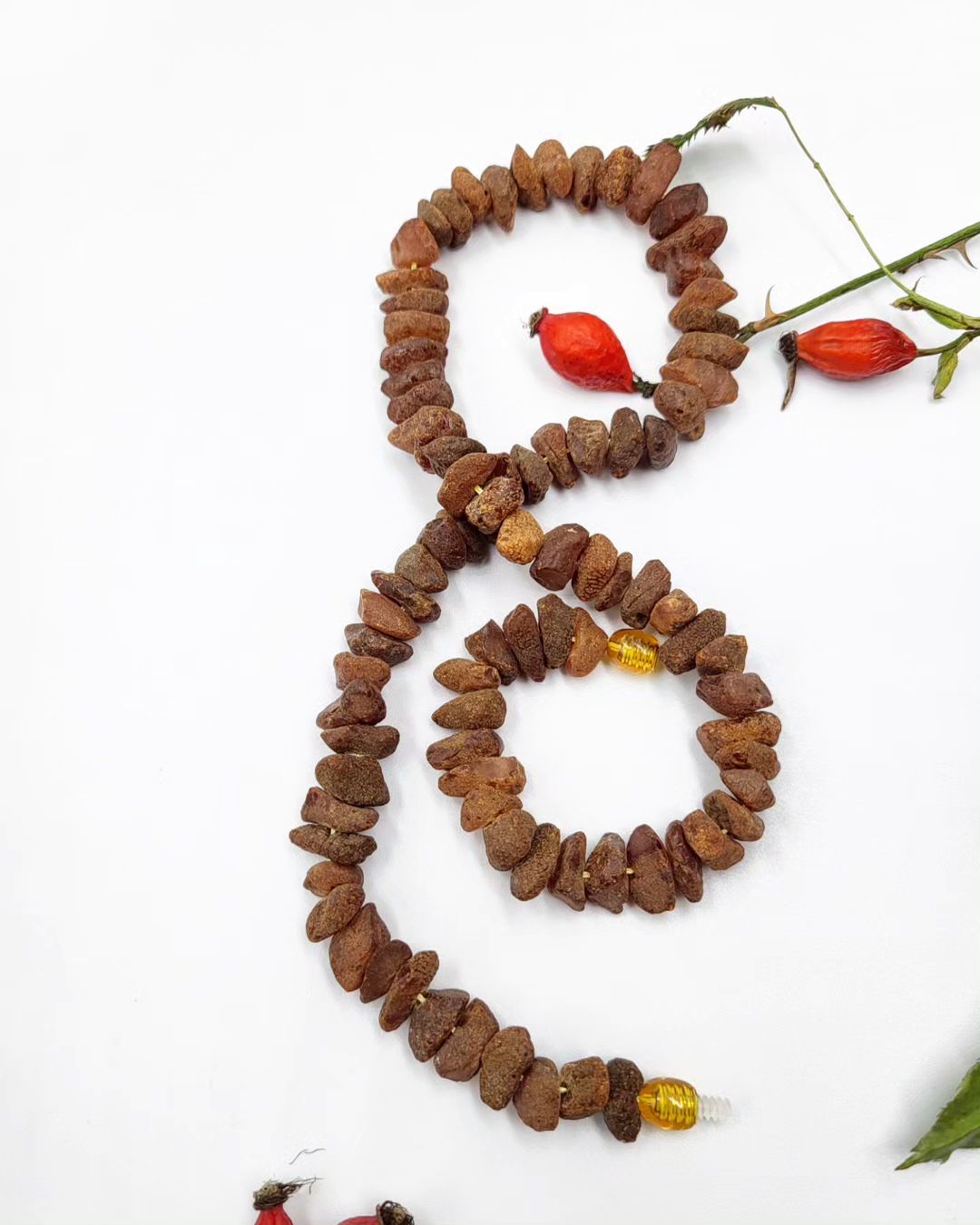 A handmade Baltic amber necklace with unpolished amber beads in varying shades of brown and a single polished yellow amber bead, displayed against a white background with rosehip berries.