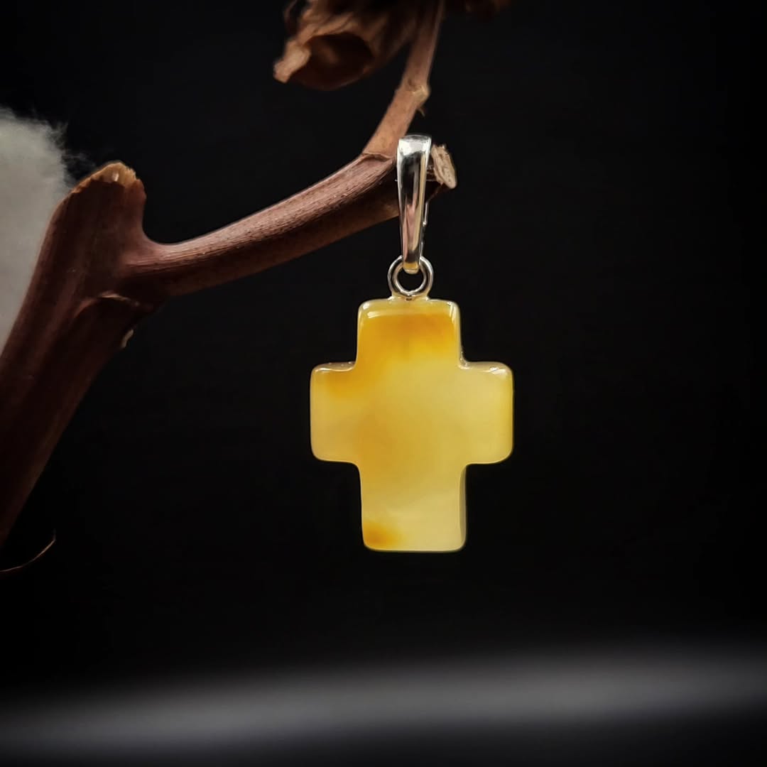 A silver-colored amber cross pendant hanging from a metal chain, displayed against a dark background with a branch.