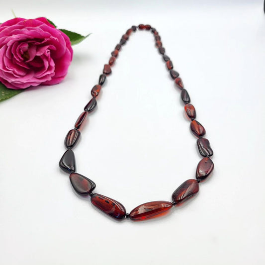 A reddish amber teardrop necklace laid out next to a pink rose. Polierte Bernsteinkette Damen dunkel Cognac – Naturbernstein Nugget Form