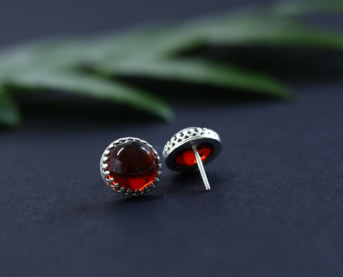 aufnahme von Tetibel Jewels Cognac Baltische Bernstein Ohrstecker (⌀12mm) in 925 Sterling Silber Kronenfassung. Die Ohrstecker liegen nebeneinander auf weißem Untergrund mit einem unscharfen roten Zweig im Hintergrund.