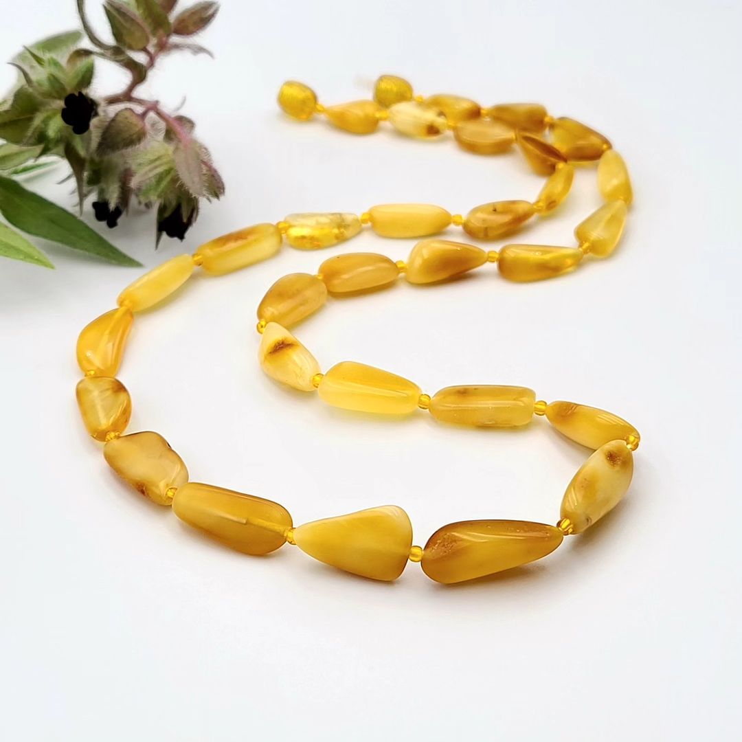 A honey-colored amber teardrop necklace with interspersed small plastic beads, displayed against a white background with a sprig of herbs to the side.