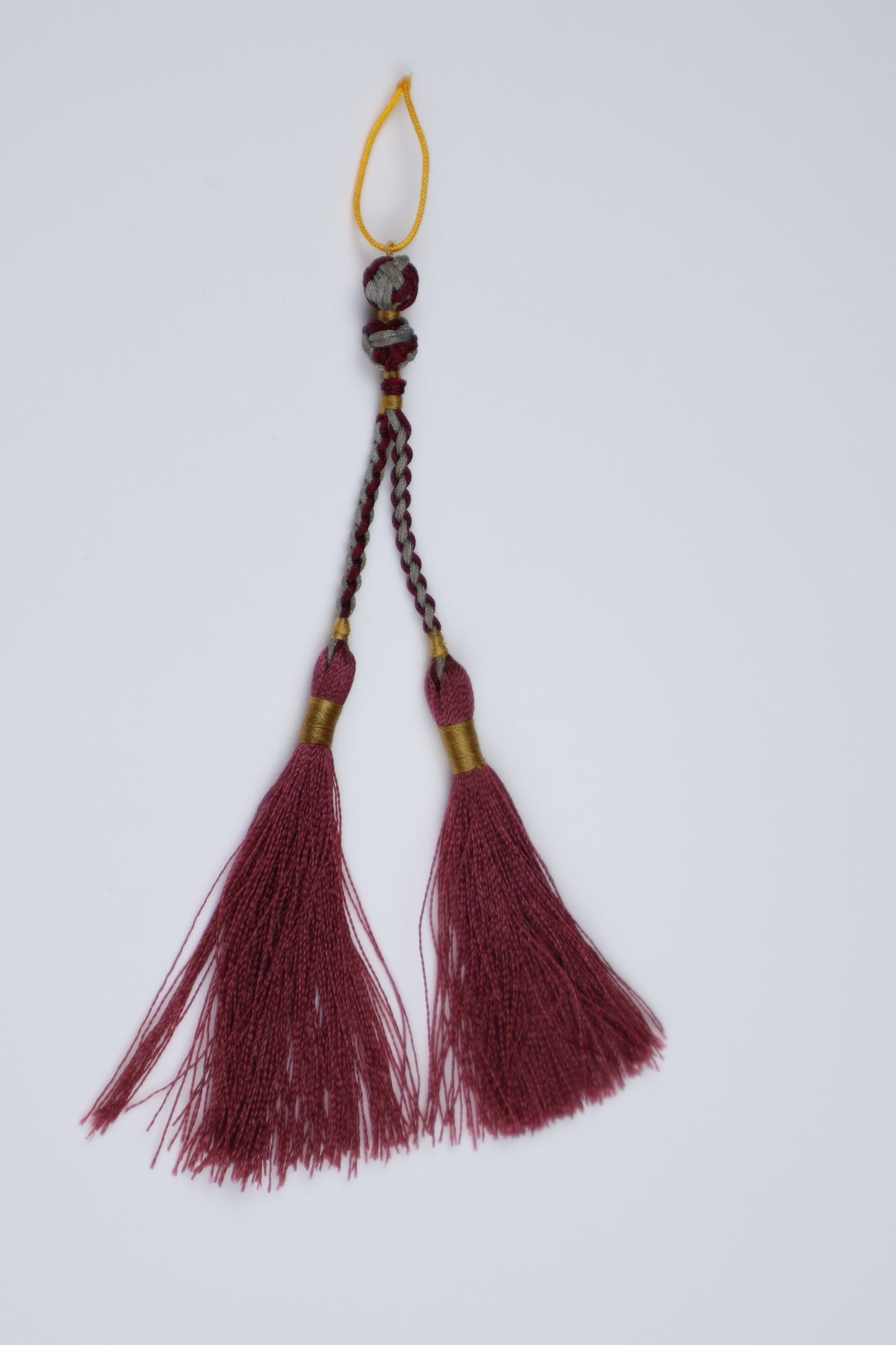 A decorative tassel ornament with two dark red fringe tassels, a braided cord, and a small yellow loop, shown on a plain light gray background.