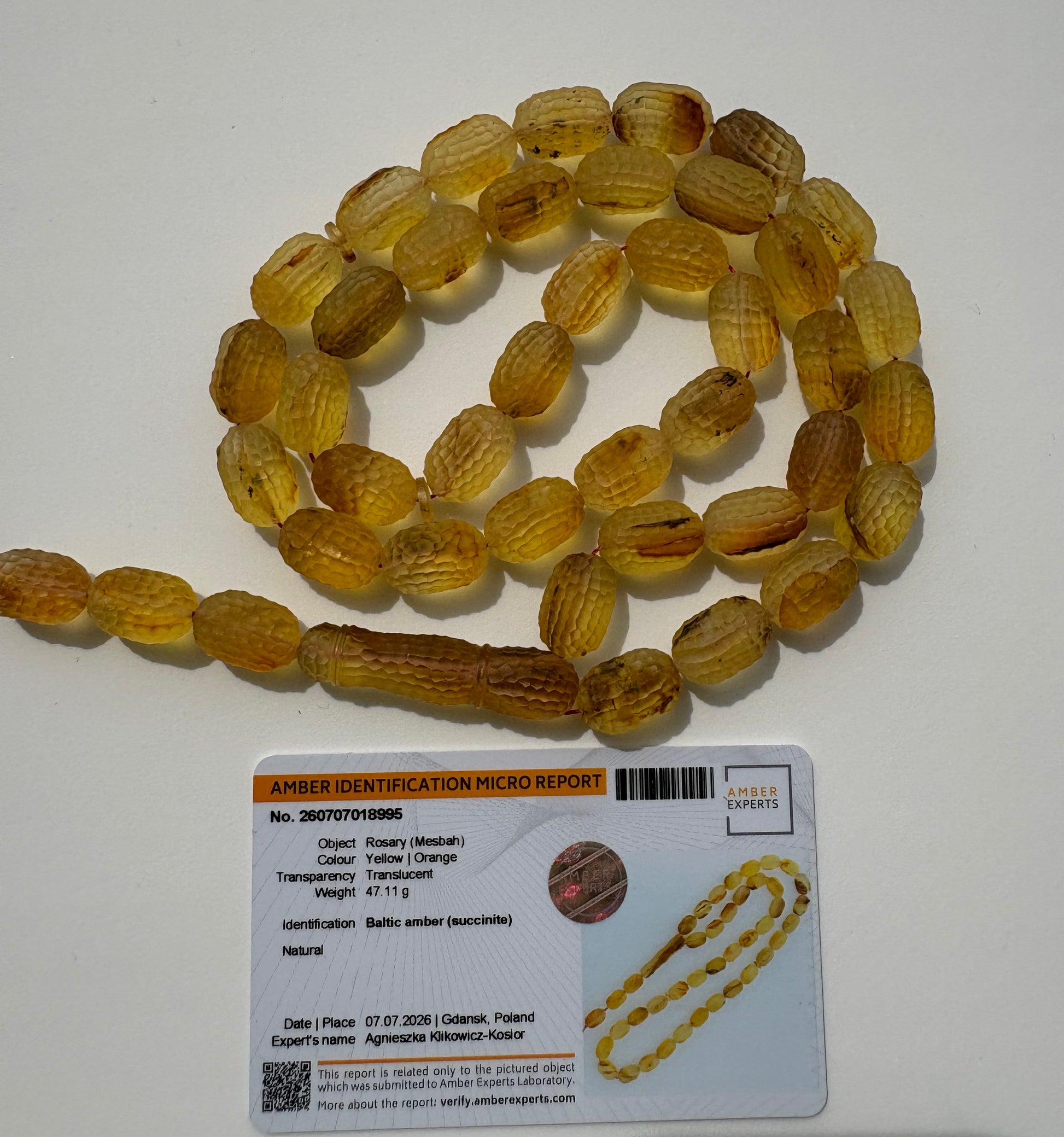 A strand of faceted, ribbed amber beads in a translucent golden-yellow hue, coiled on a white surface next to a product identification card.