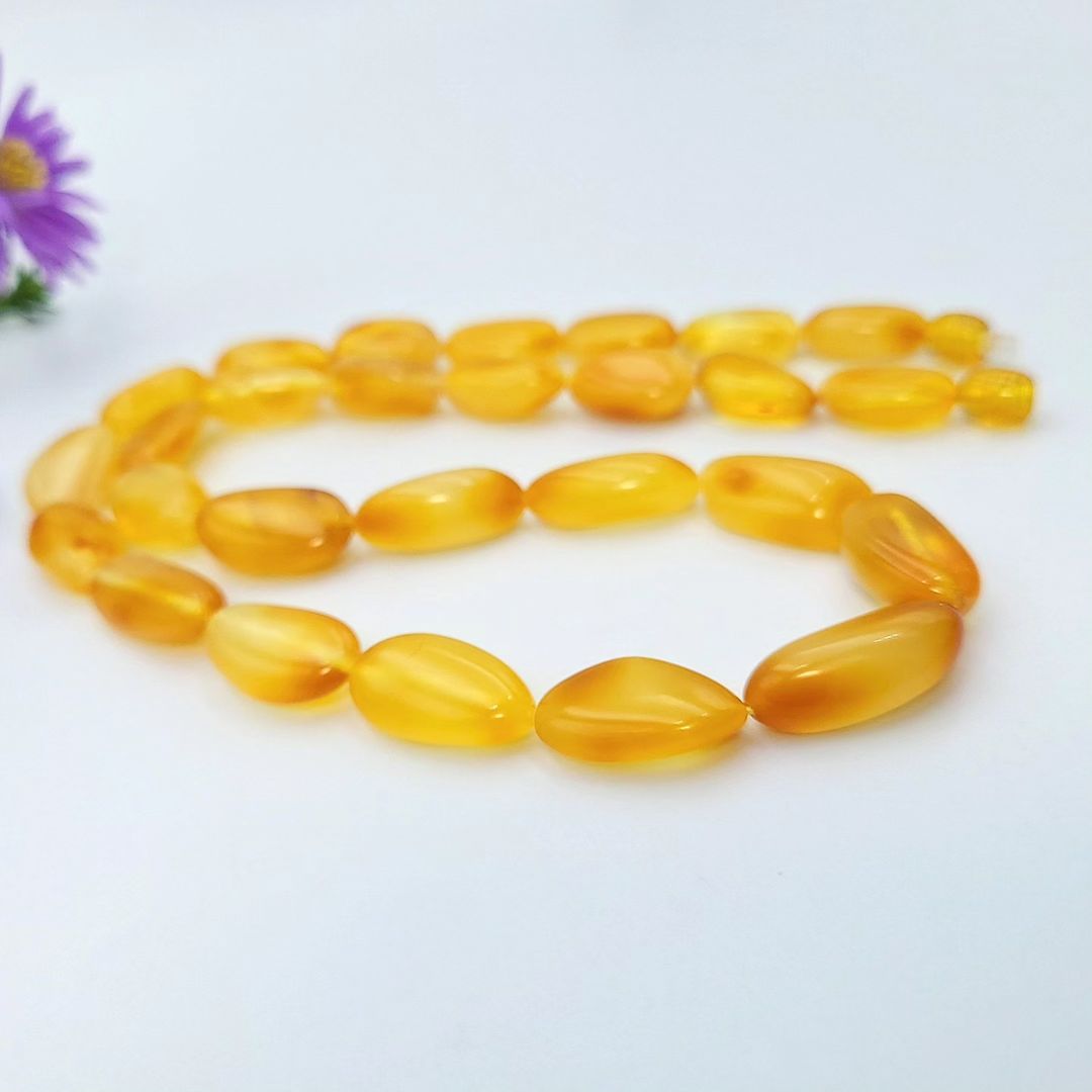 A full view of a bright honey-yellow amber necklace loosely coiled in a spiral on a white surface. The polished, irregular, tumbled beads are graduated, getting smaller towards the ends. A purple aster flower is visible in the upper left corner.