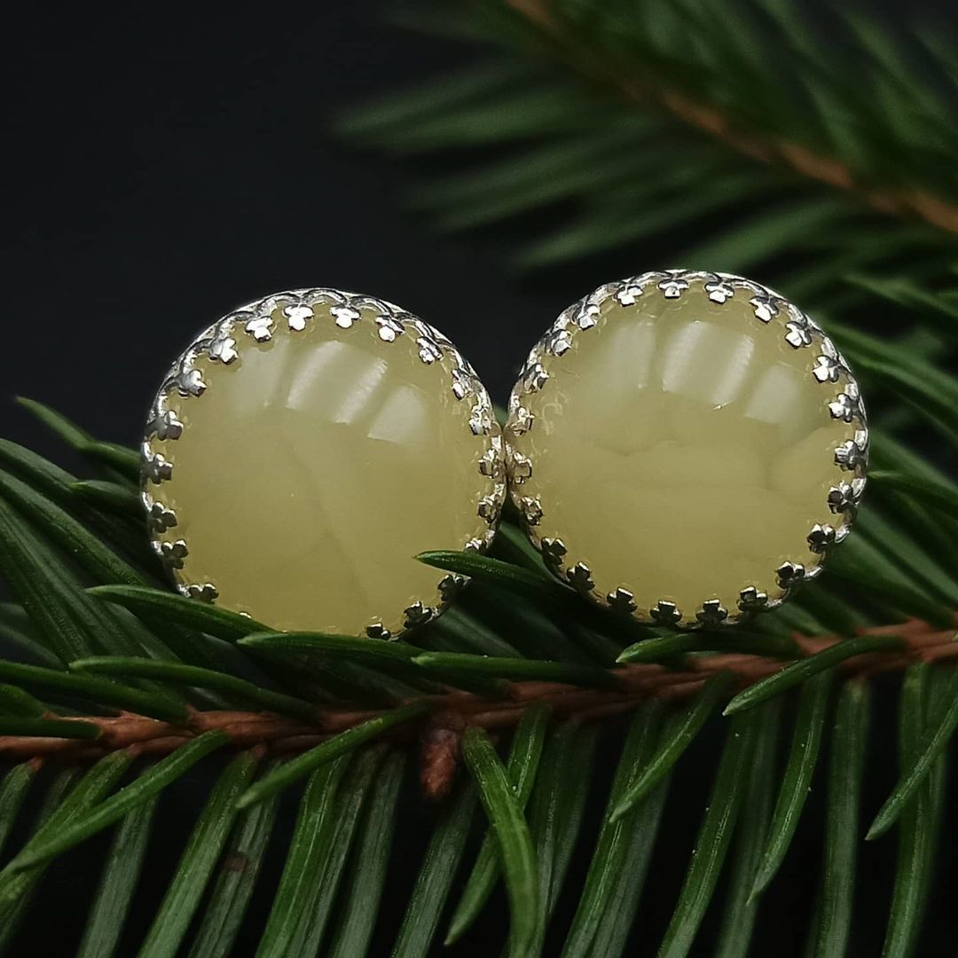 Tetibel Jewels Butterscotch Baltische Bernstein Ohrstecker (⌀12mm) in Seitenansicht, zeigt den Bernstein in der 925 Sterling Silberfassung und den Ohrsteckerstift. Liegt auf dunklem Untergrund mit einem verschwommenen Blumenstiel.