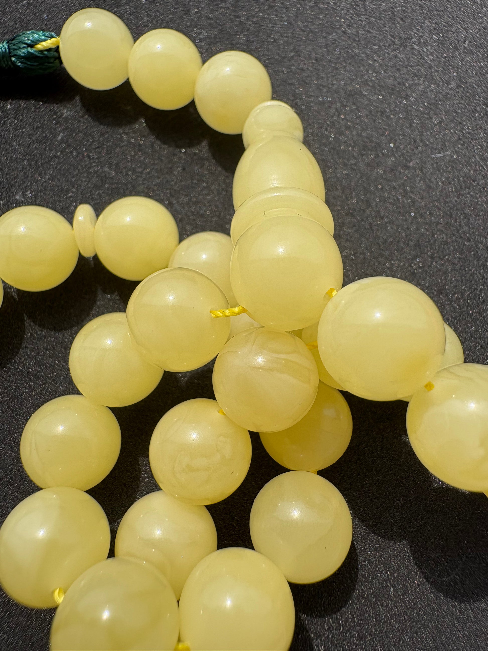 A close-up of Tetibel pale yellow translucent round amber rosary on a dark textured surface.