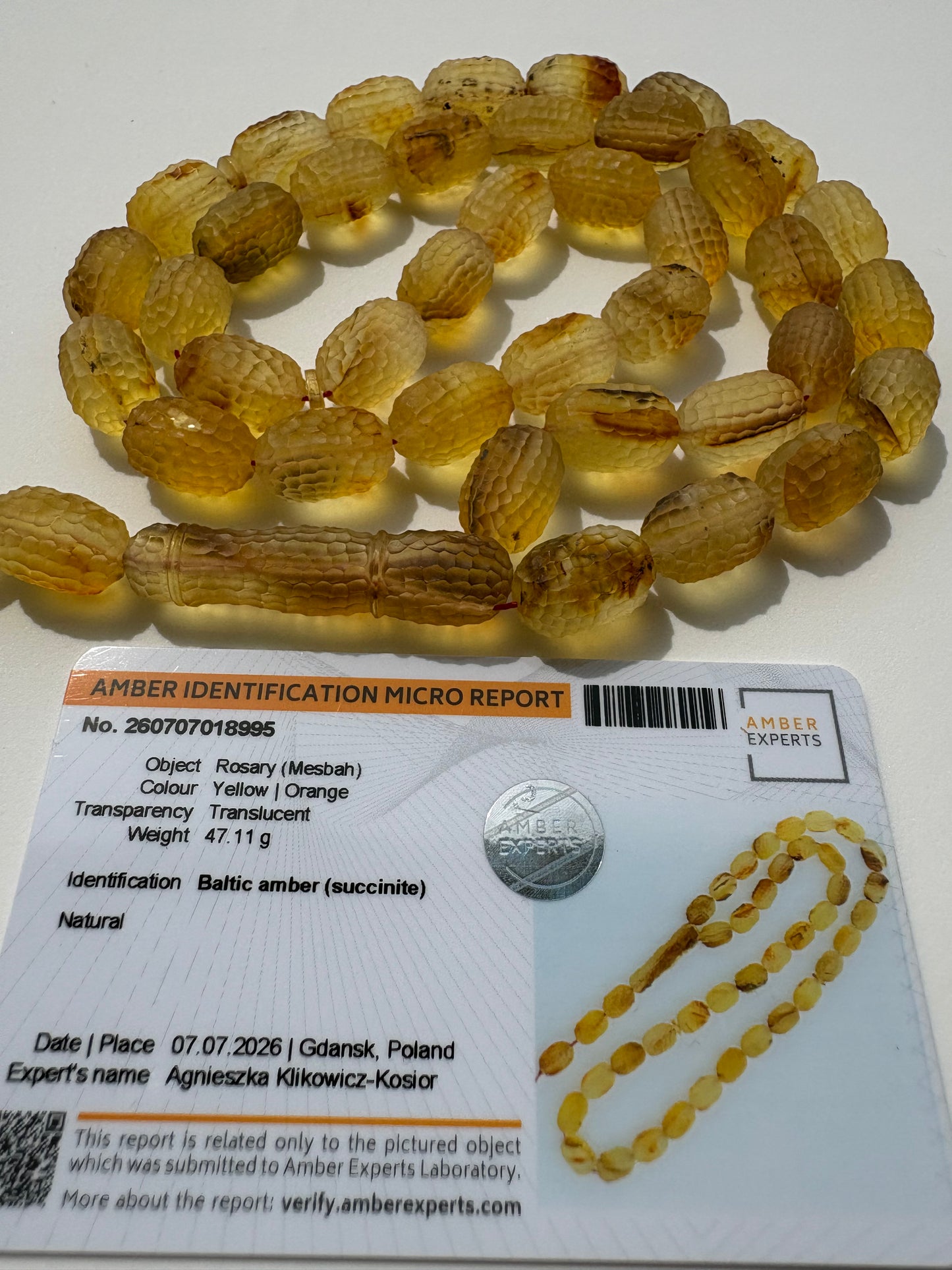 A strand of amber beads with a detailed identification card next to it, photographed on a white surface. The beads are translucent golden-yellow with natural brown inclusions.