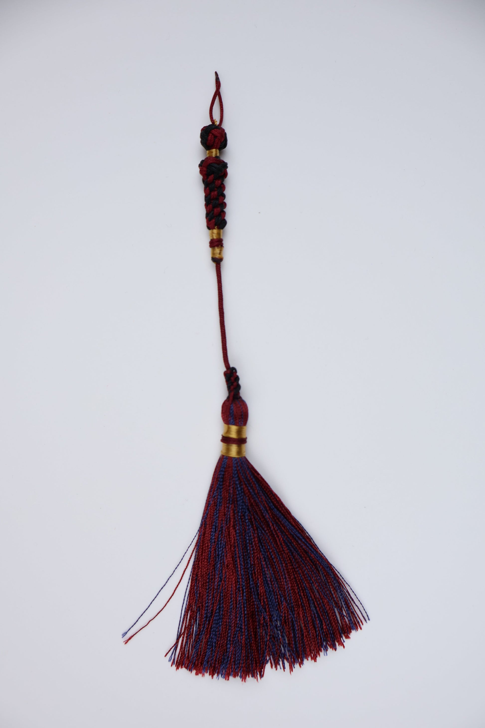 A decorative tassel with a dark red and navy blue fringed end, a gold-colored metal collar, and a knotted cord section with small gold beads, shown on a plain white background.