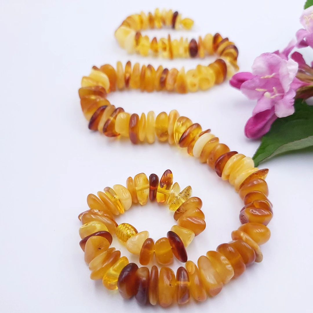 A cascade-style amber necklace with chips in varying shades of yellow and brown, displayed against a white background with pink flowers to the side.