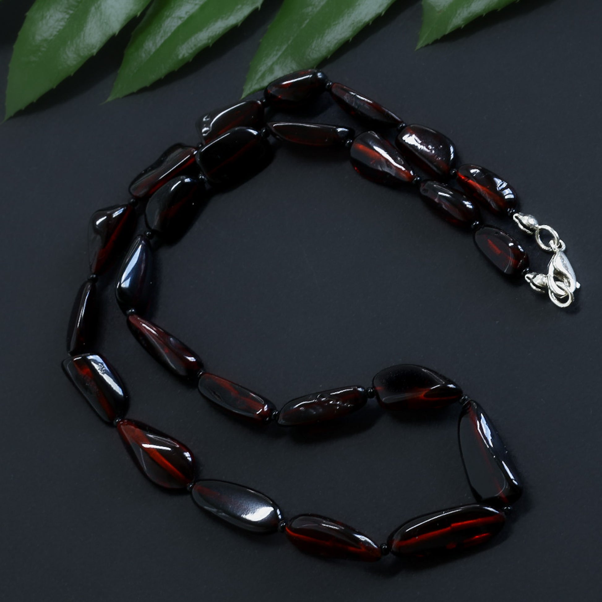 Beaded necklace with a silver clasp on a dark surface with green leaves in the background Polierte Bernsteinkette Damen dunkel Cognac – Naturbernstein Nugget Form