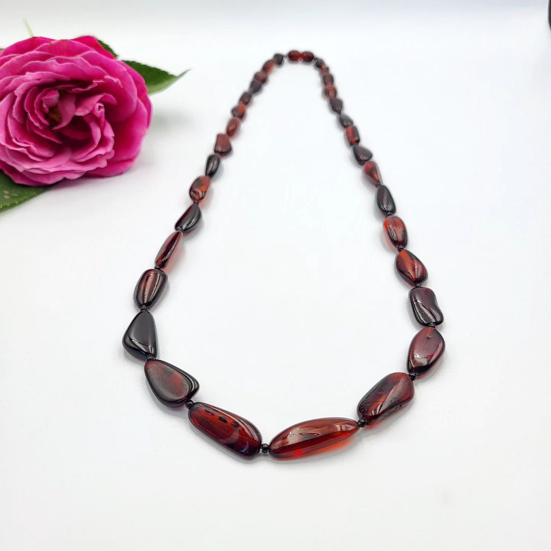 A reddish amber teardrop necklace laid out next to a pink rose. Polierte Bernsteinkette Damen dunkel Cognac – Naturbernstein Nugget Form