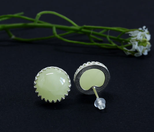 Nahaufnahme von Tetibel Jewels Butterscotch Baltische Bernstein Ohrstecker (⌀12mm) in 925 Sterling Silber Kronenfassung. Die Ohrstecker liegen nebeneinander auf dunklem Untergrund mit einem unscharfen Blumenstiel im Hintergrund.