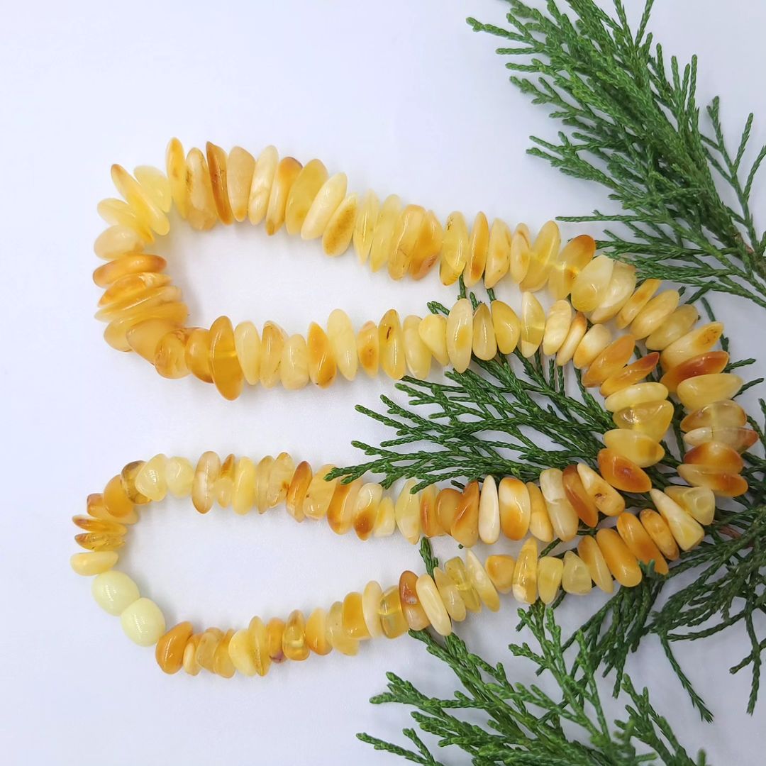Nahaufnahme der Tetibel echten baltischen Rohbernstein-Splitterkette in Butterscotch und Lemon. Auf weißem Hintergrund mit grünen Zweigen in geschwungener Form. Zeigt die unpolierten Nuggets und den Kunststoff-Schraubverschluss.