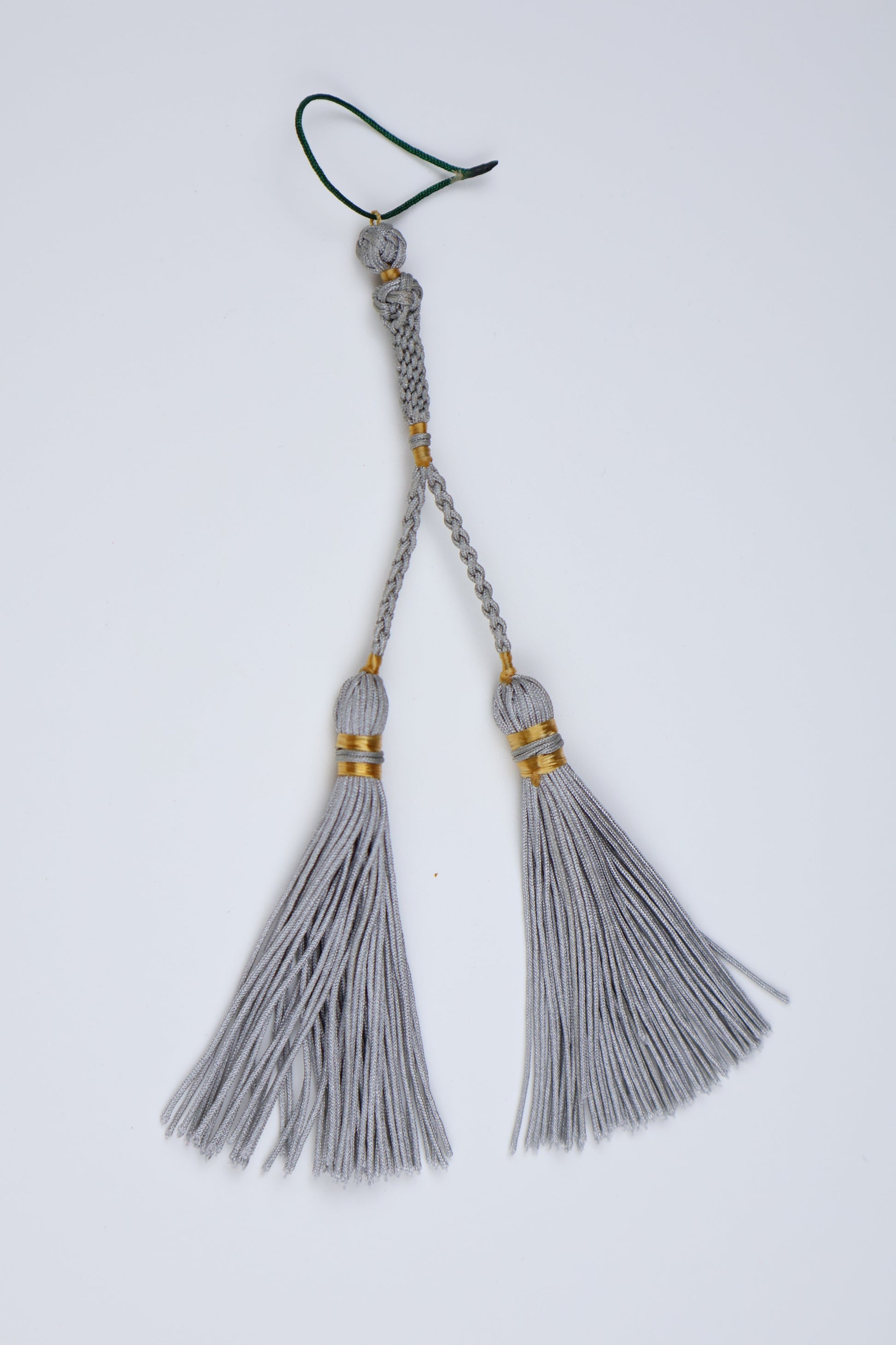 A pair of light gray decorative tassels with gold accent bands and a braided cord leading to a small green loop, photographed on a plain white background.